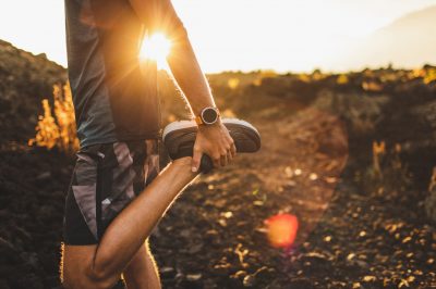 Male runner stretching leg and feet and preparing for running outdoors. Smart watch or fitness tracker on hand. Beautiful sun light on background. Active and healthy lifestyle concept.